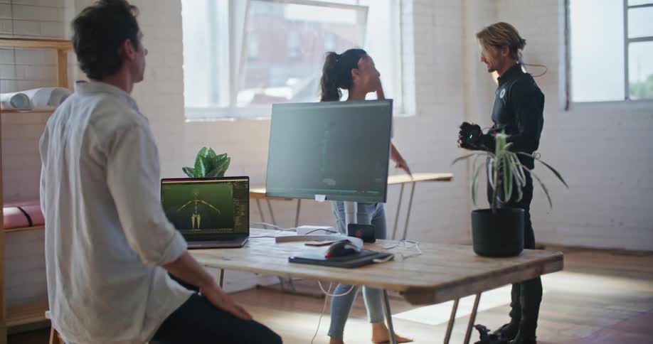 A man is sitting at a desk with computers and a man wearing virtual reality devices is practicing movements with a woman