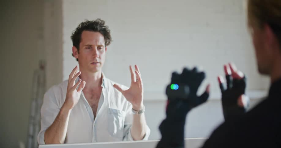 A man has interactive computer devices on his hands and another man looks at one and shows him hand movements