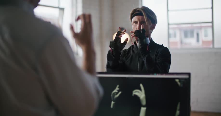 A man has interactive computer virtual reality devices on his hands and his hand movements are seen on a computer monitor