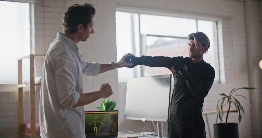 A man wearing virtual reality equipment moves his hands and arms as instructed by another man who makes adjustments and looks at movements on a computer