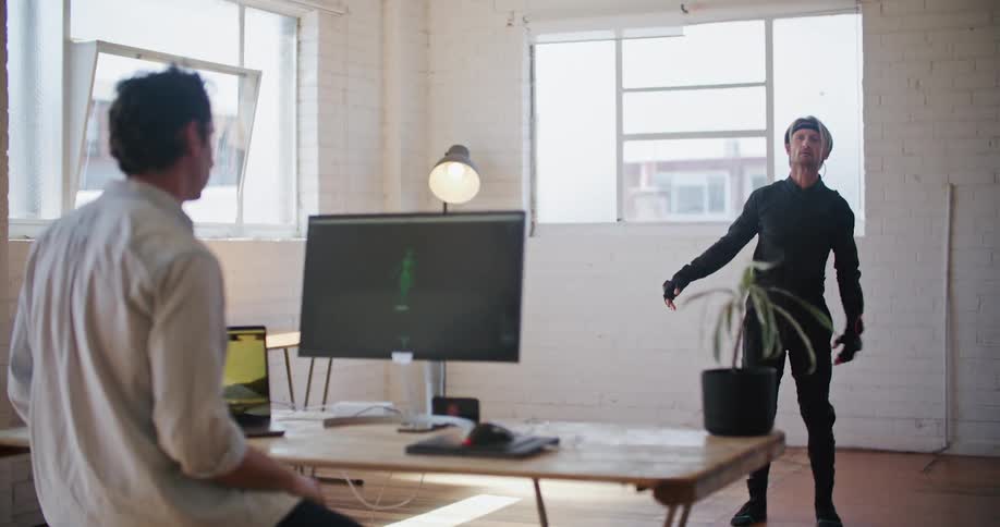 A man wearing virtual reality devices is waving his arms in the air as another man watches his movements on a computer monitor