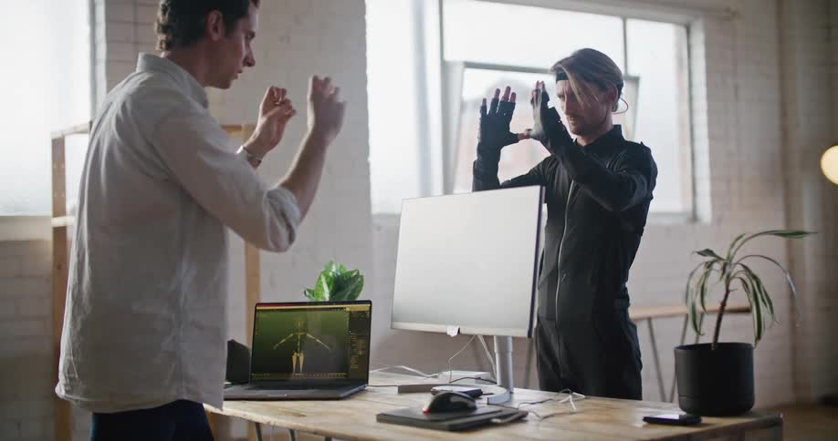 A man wearing interactive computer virtual reality devices is speaking with a man standing behind a computer and imitating his hand movements