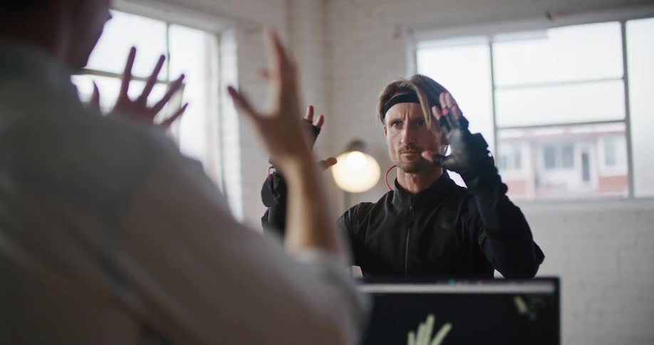 A man has virtual reality devices on his hands and he is imitating another persons hand movements and they are shown on a screen