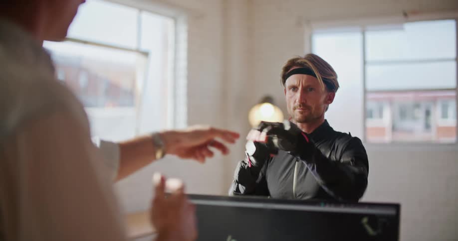 A man has interactive computer virtual reality devices on his hands and is moving his hands as he speaks to a man sitting behind a computer