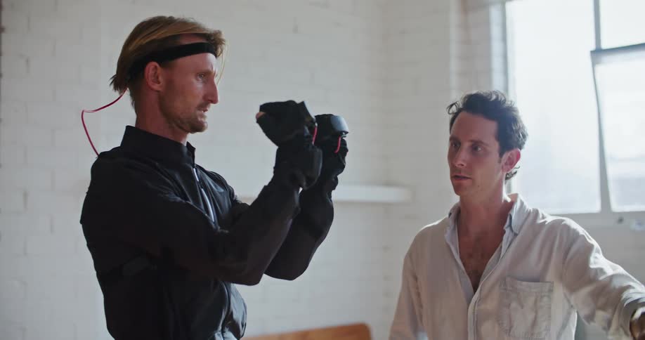 A man has interactive computer devices on his hands and another man shows him hand movements