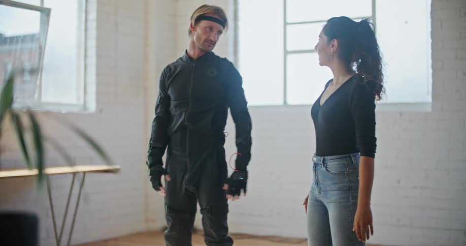 A man wearing interactive computer virtual reality devices is doing martial arts movements and is standing next to a woman who is showing him movements