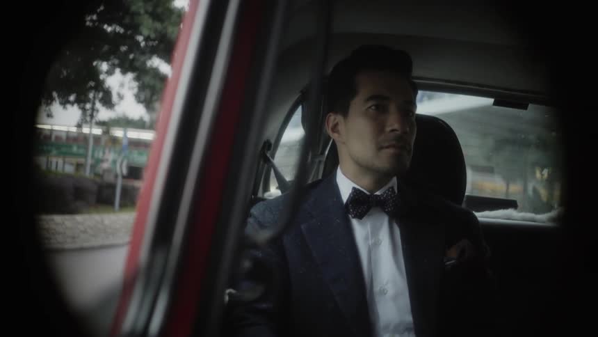 A man in a suit sits in a car and smiles as he is driven through a city