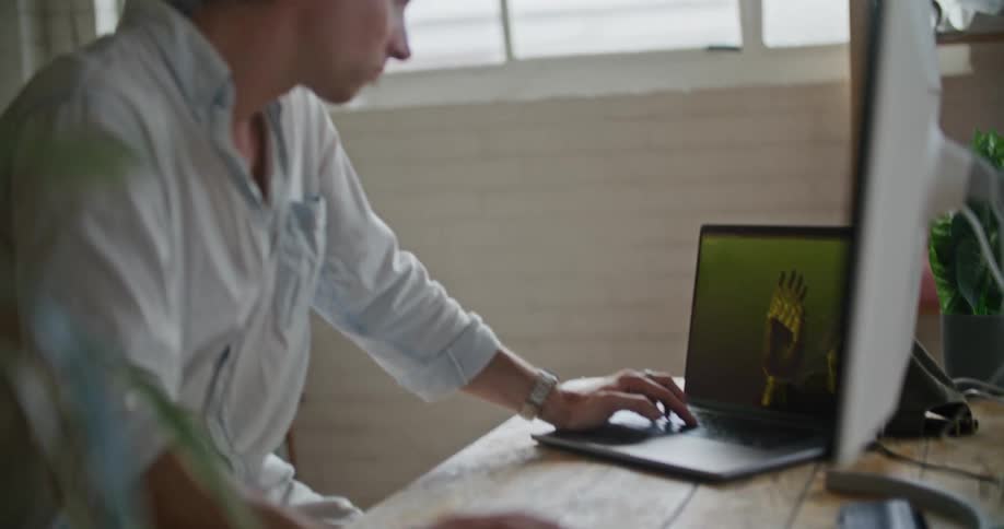 A man is sitting at a desk looking at a large computer monitor and then over at a laptop computer