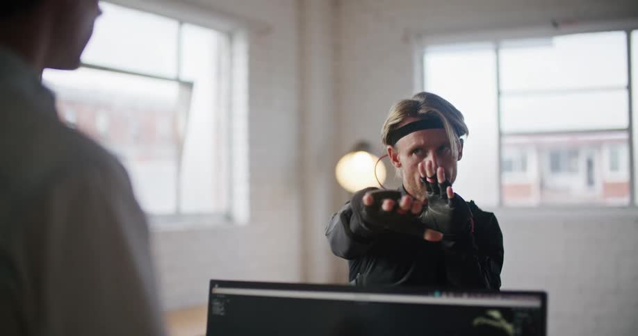 A man has virtual reality devices on his hands and his hand movements are seen on a computer monitor as he talks to another man