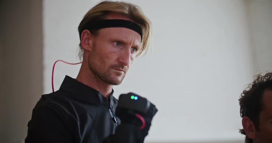A man wearing interactive computer virtual reality devices is moving his hand and looking at it as another man sits near him