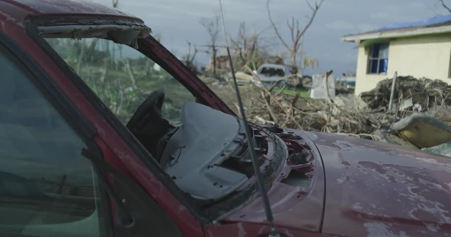 Wrecked vehicles sit in yard of house with roof tarp among plant and other debris