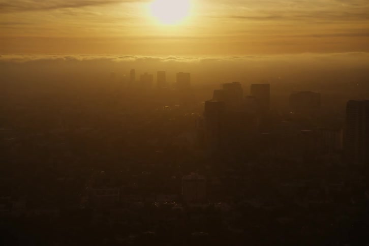 Silhouettes of buildings can be seen as the sun shines brightly in a hazy orange sky