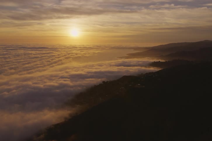 The sun is shining brightly above clouds and fog over the coastline next to mountains