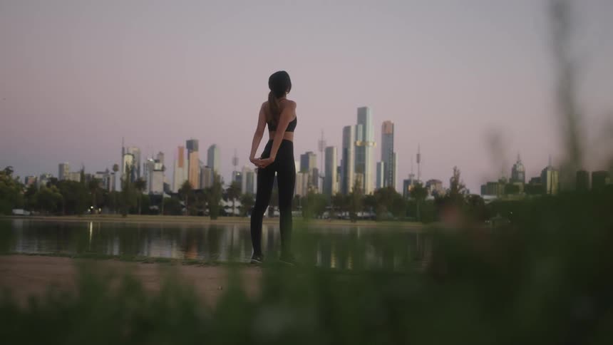 A woman stands on the shore and stretches with a downtown city skyline in the distance
