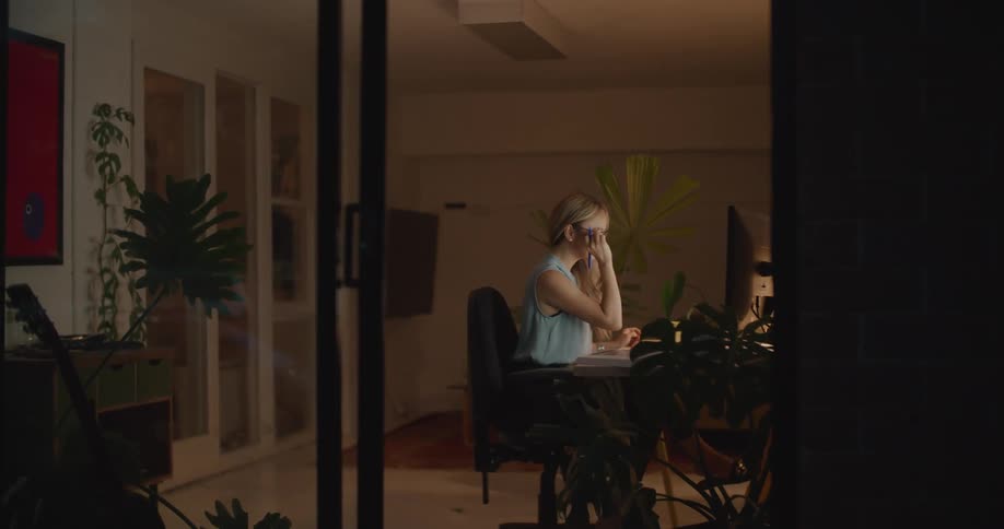 Woman with brown wavy hair glasses and blue blouse sits at desk working on computer in room at night
