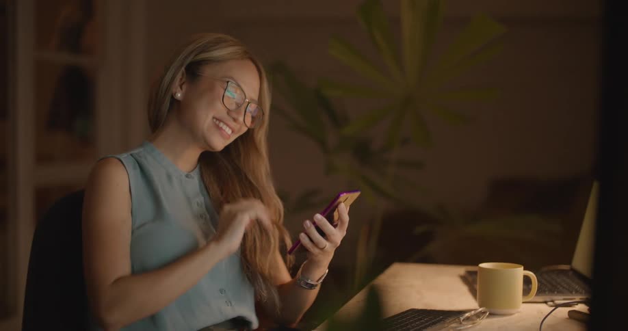 Woman with brown wavy hair glasses and blue blouse sits at desk using smartphone and smiling in the office