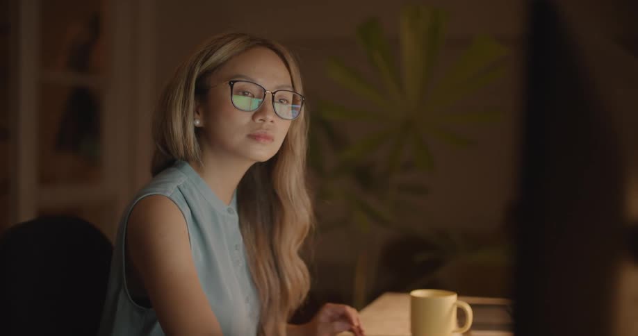 A young woman in an office sits at her desk and looks at her computer and writes something down