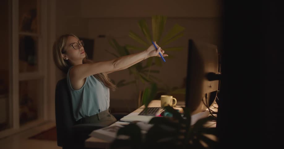 A woman sitting in her office stretches and then types before taking off her glasses