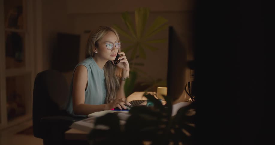 A woman talking on her cell phone and working on a computer at night