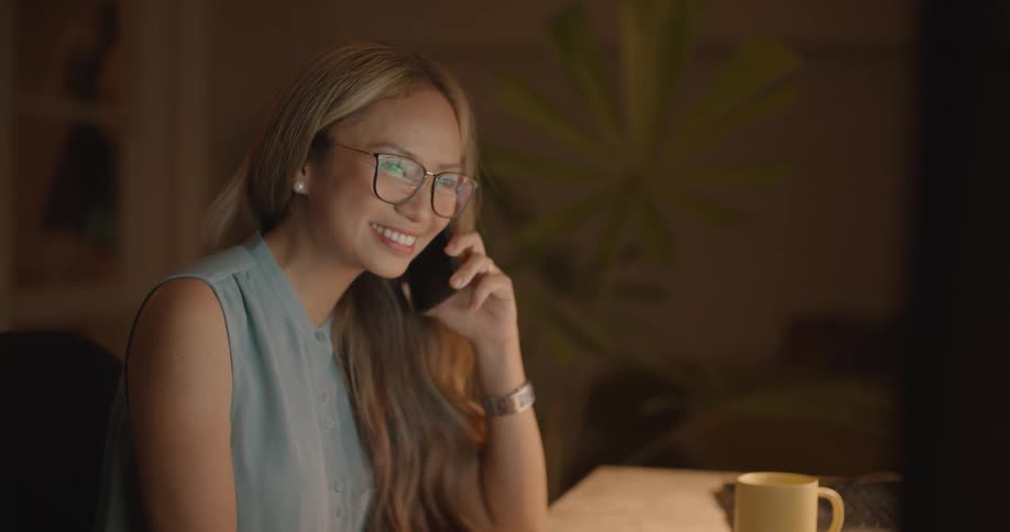 Woman with long brown wavy hair glasses and blue blouse sits at desk and ends a call on smartphone