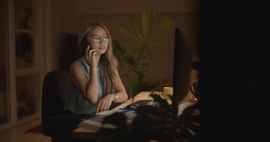 Woman with long brown hair glasses and blue blouse talks on smartphone while working on computer in room