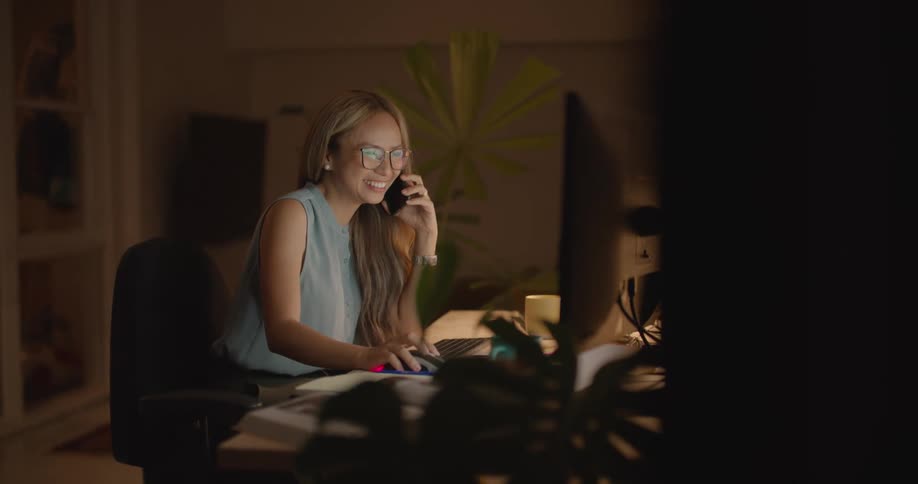 Young asian woman talking on cell phone while working in office at night