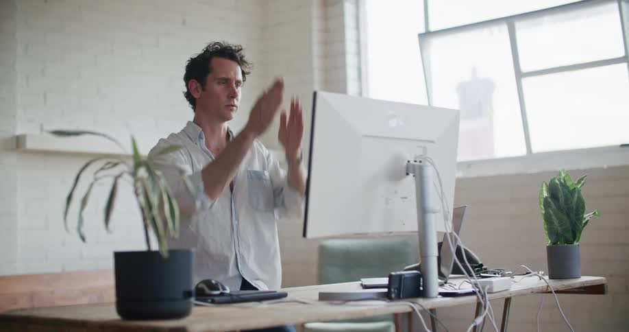 A man is sitting at a desk behind a computer and making hand motions as he speaks to someone