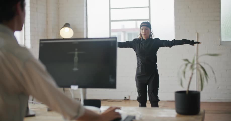 A woman with virtual reality equipment on stands in an office and moves her arms as a man tells her to as he looks at an image on a computer