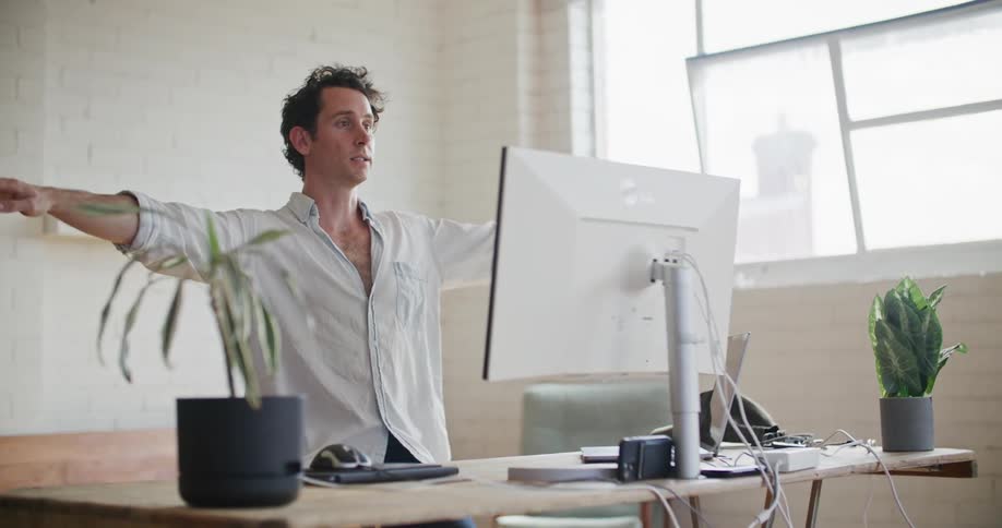 A man makes a face then stretches his arms out while he talks on a video call on a computer