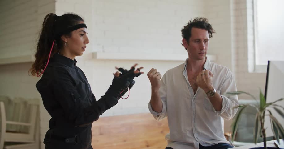 A woman with virtual reality equipment on stands next to a man in an office and they move their hands as he looks at a computer