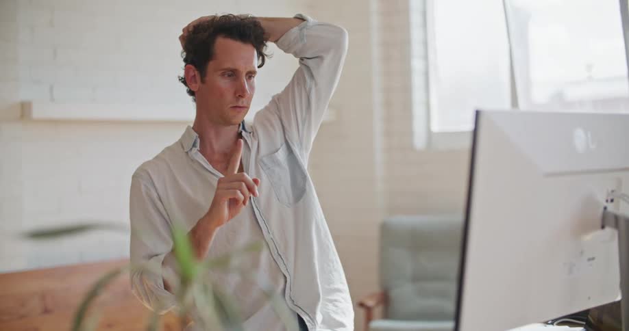 A man in an office holds his finger up to his computer as he video chats and runs his fingers threw his hair and scratches his head
