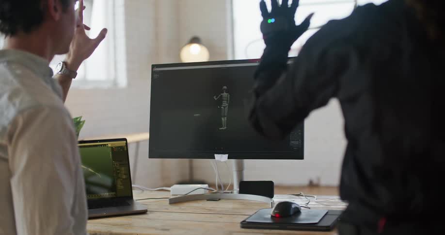 A woman with virtual reality equipment on stands next to a man in an office and they move their hands as they watch a robot on a computer monitor