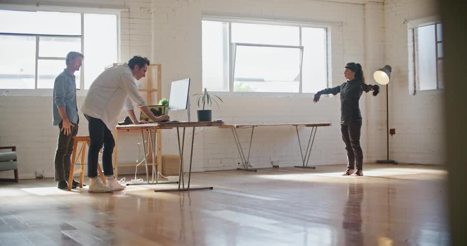 A woman with virtual reality equipment on stands in an office an mimics a mans actions as another man looks at a computer