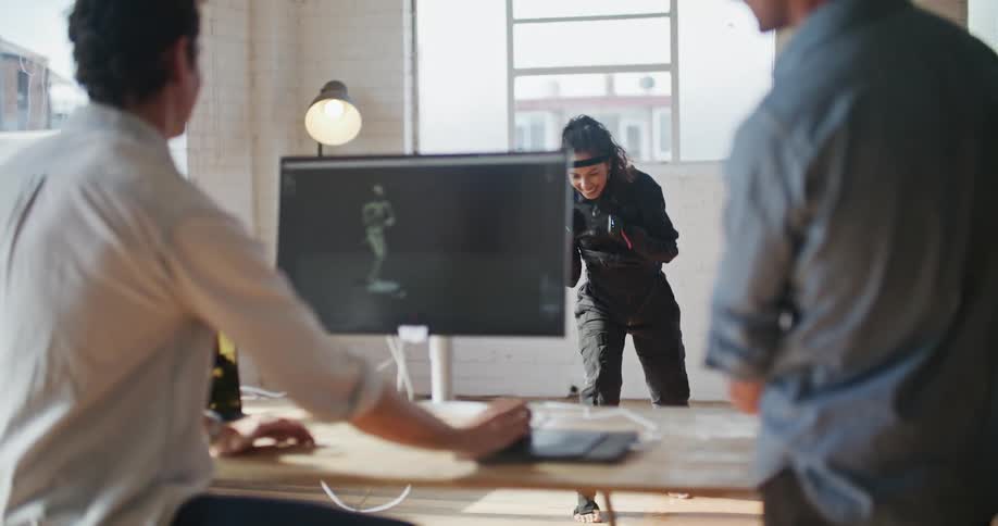 Smiling men watch woman wearing a motion capture suit punch the air as the nearby screen show animated figure moving through the same motions