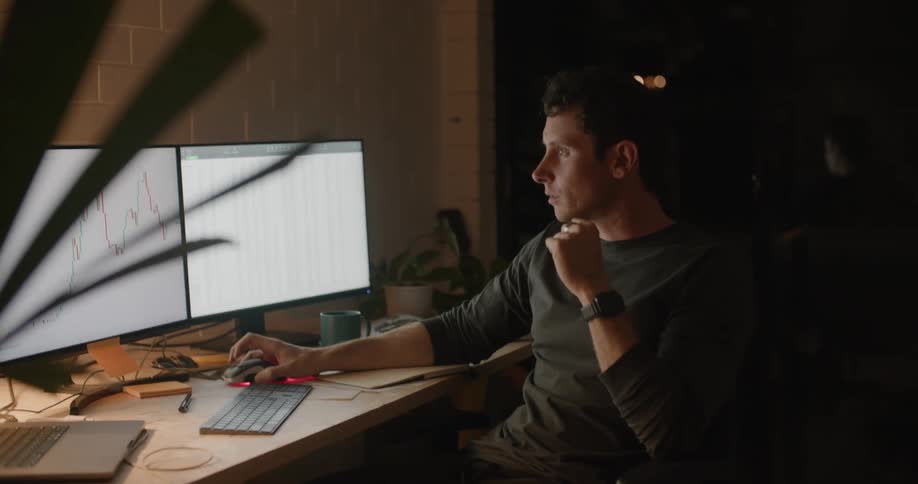 Man with gray sweatshirt sitting at desk works on computers and writes down in notebook