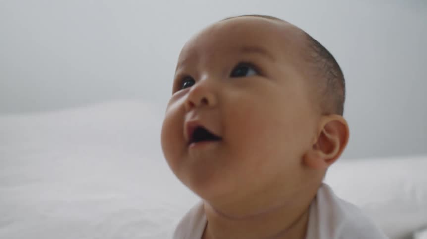 A baby sits on a bed as someone plays with him and puts a sheet over his head and pulls it off