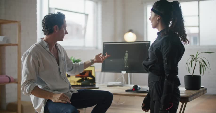 Woman wearing motion capture suit stands talking to man giving instruction next to computers showing animation