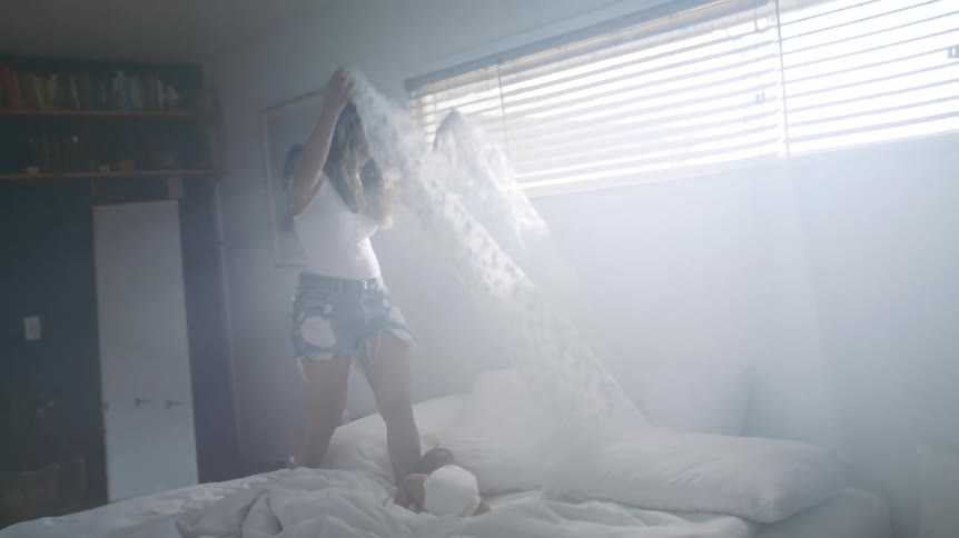 A woman stands on her bed and plays with her young baby by throwing a sheet over him and removing it