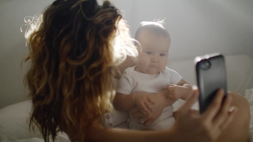 Mother with brown wavy hair holds baby and makes video call on smartphone
