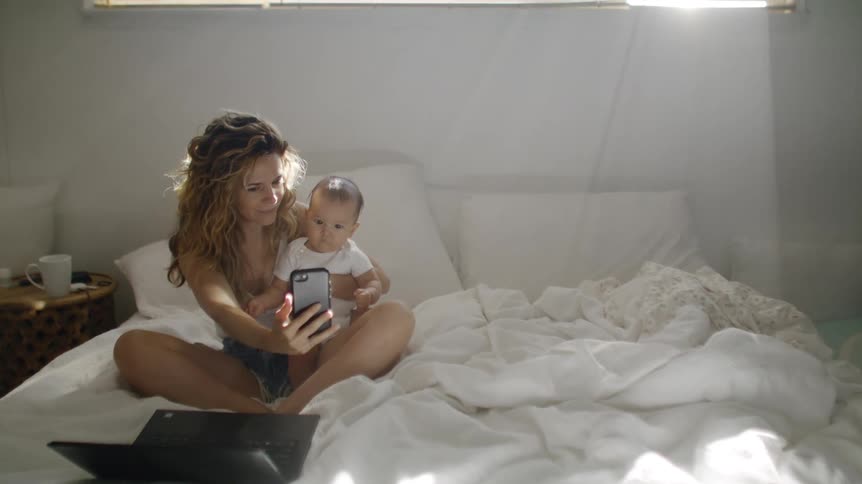 Mother with brown wavy hair white sleeveless shirt and shorts sits on bed holding baby son and taking selfie in bedroom