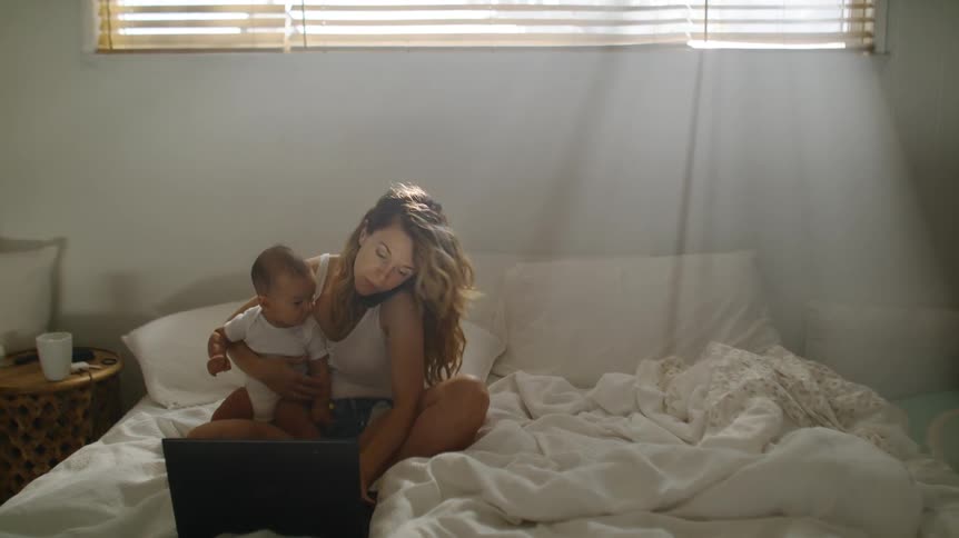 Mother holding her baby sitting on bed working on laptop while talking on cell phone