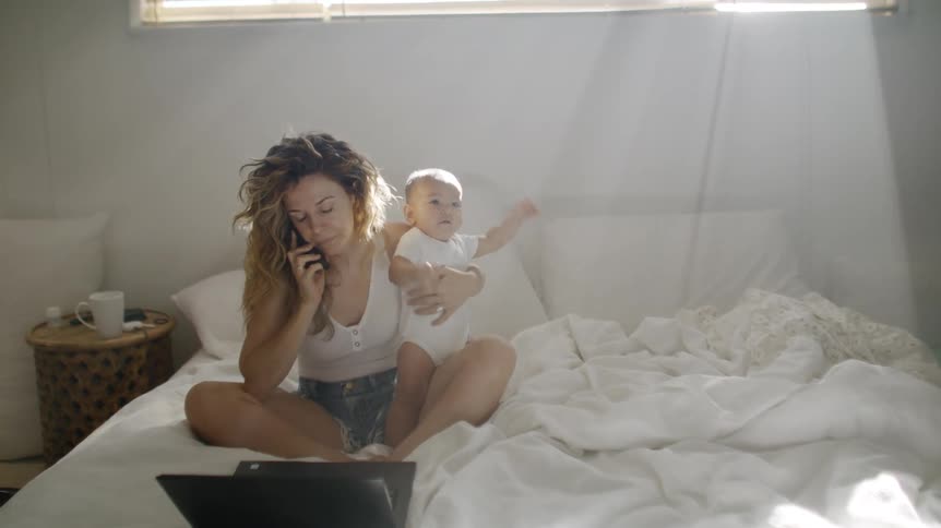 Mother sitting in the bed using her laptop and talking to the phone while holding her baby