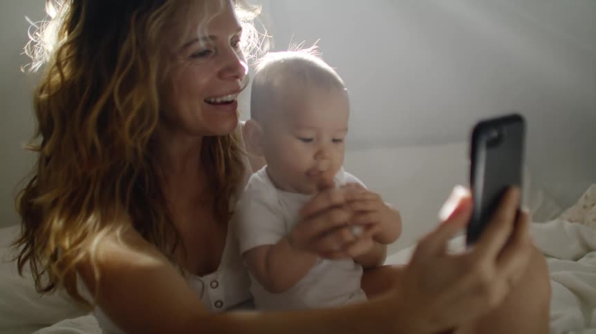 Mother sitting on bed taking a selfie with baby on cell phone