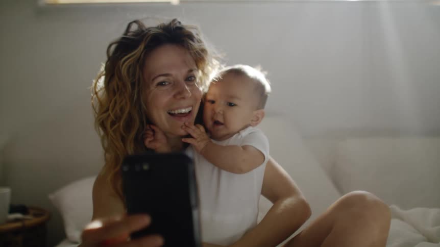 Mother sitting on the bed holding her baby and taking a selfie with her smartphone