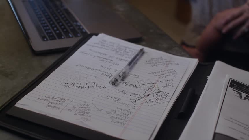 A woman stands swaying back and forth with a baby on her hip working on a laptop and taking notes