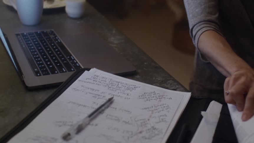 Woman with brown wavy hair and gray sweatshirt holds baby and writes down in notebook quickly