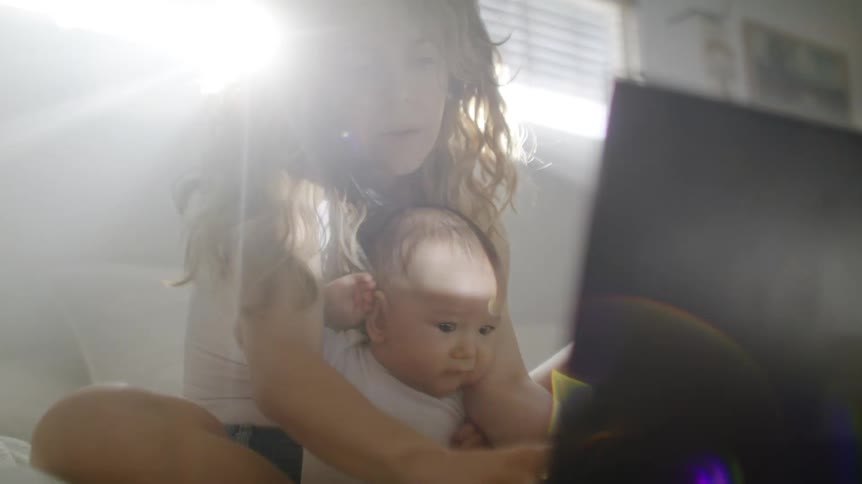 Woman sitting in the bed being bothered by her baby on her lap while she is talking to the phone and typing on her laptop