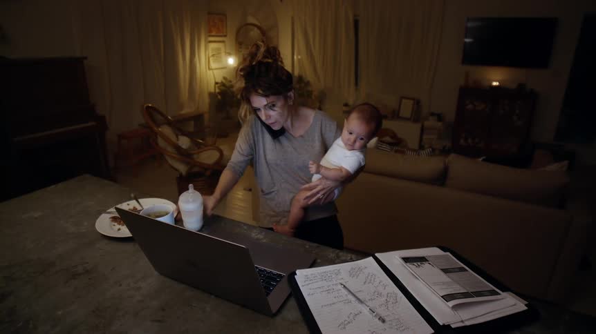 Woman standing and talking to the phone while trying to feed her baby in front of her laptop and a notebook