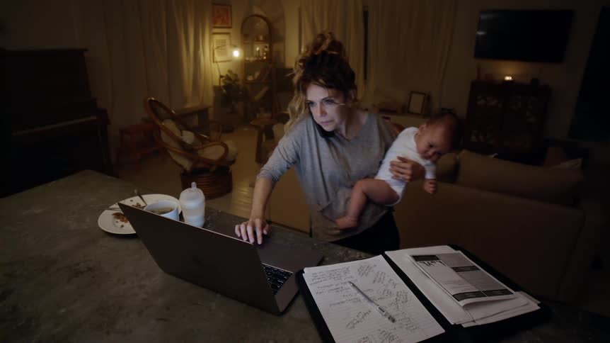 Woman stands talking on the phone while working on a computer before taking time to feed her baby