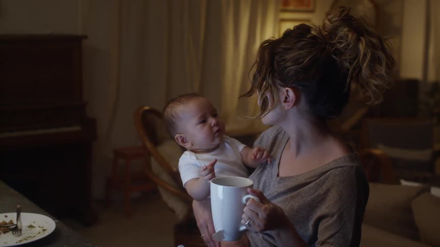 Mother sits at table near piano drinking from large cup and holding baby in one arm